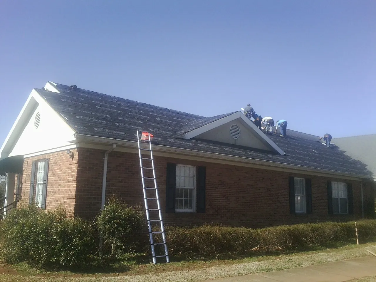 Crew tearing shingles off a brick ranch roof with a ladder set — Metal Roof Repair work by our crew in Fort Loramie