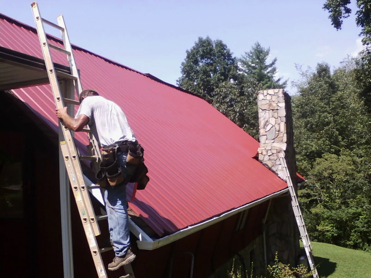 Roofer on a ladder beside a red metal roof and stone chimney — Soffit & Fascia Repair by Roofing Brothers in Fort Loramie